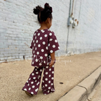 A toddler wearing a burgundy corduroy two-piece outfit with white polka dots, walking on a concrete path against a white brick wall. A 'GTRs Boutique' watermark is visible across the center.