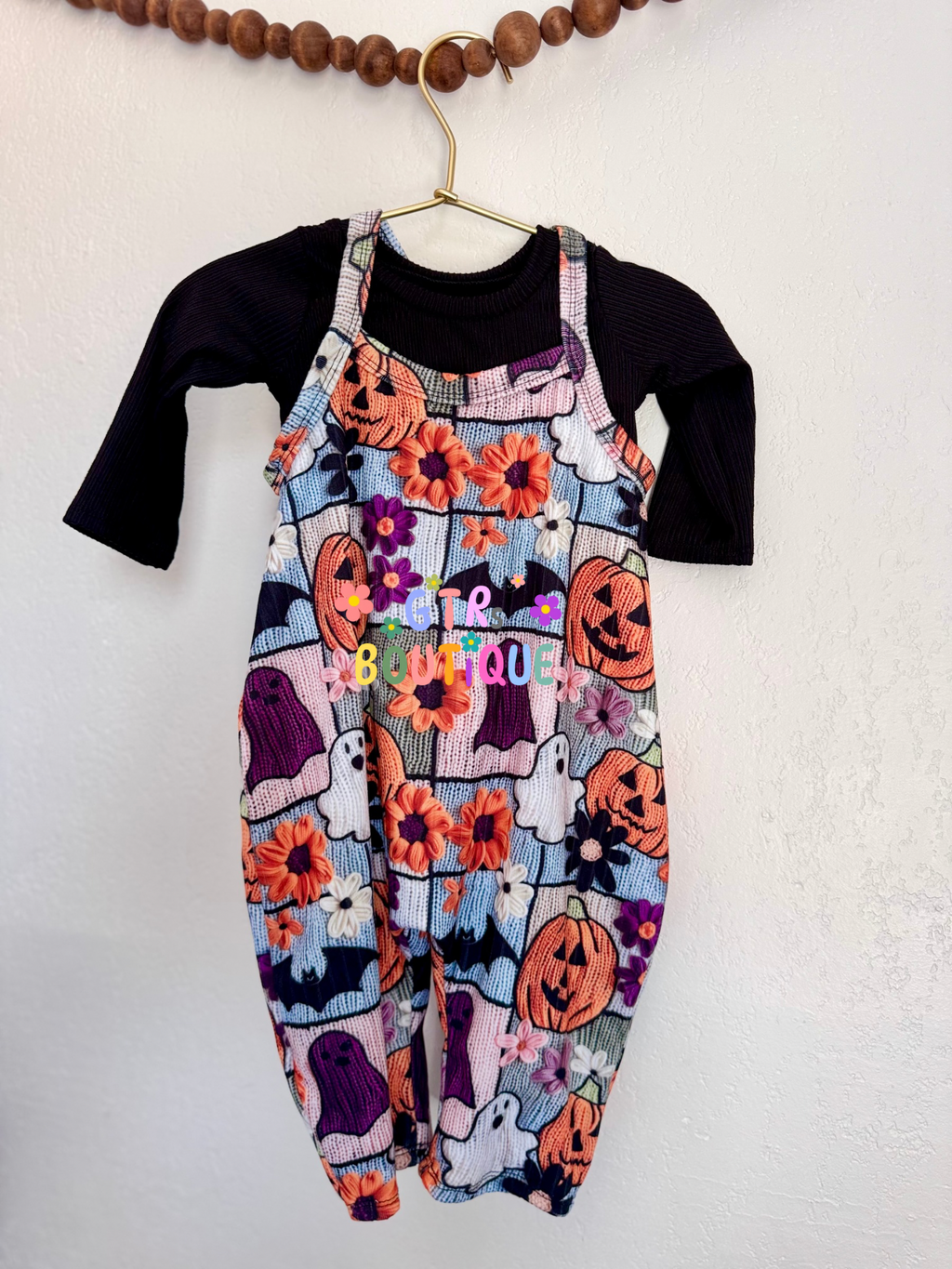 A toddler's Halloween-themed jumpsuit featuring a colorful patchwork print of jack-o'-lanterns, ghosts, pumpkins, and flowers, worn over a black long-sleeve shirt and hanging on a gold hanger against a white wall.